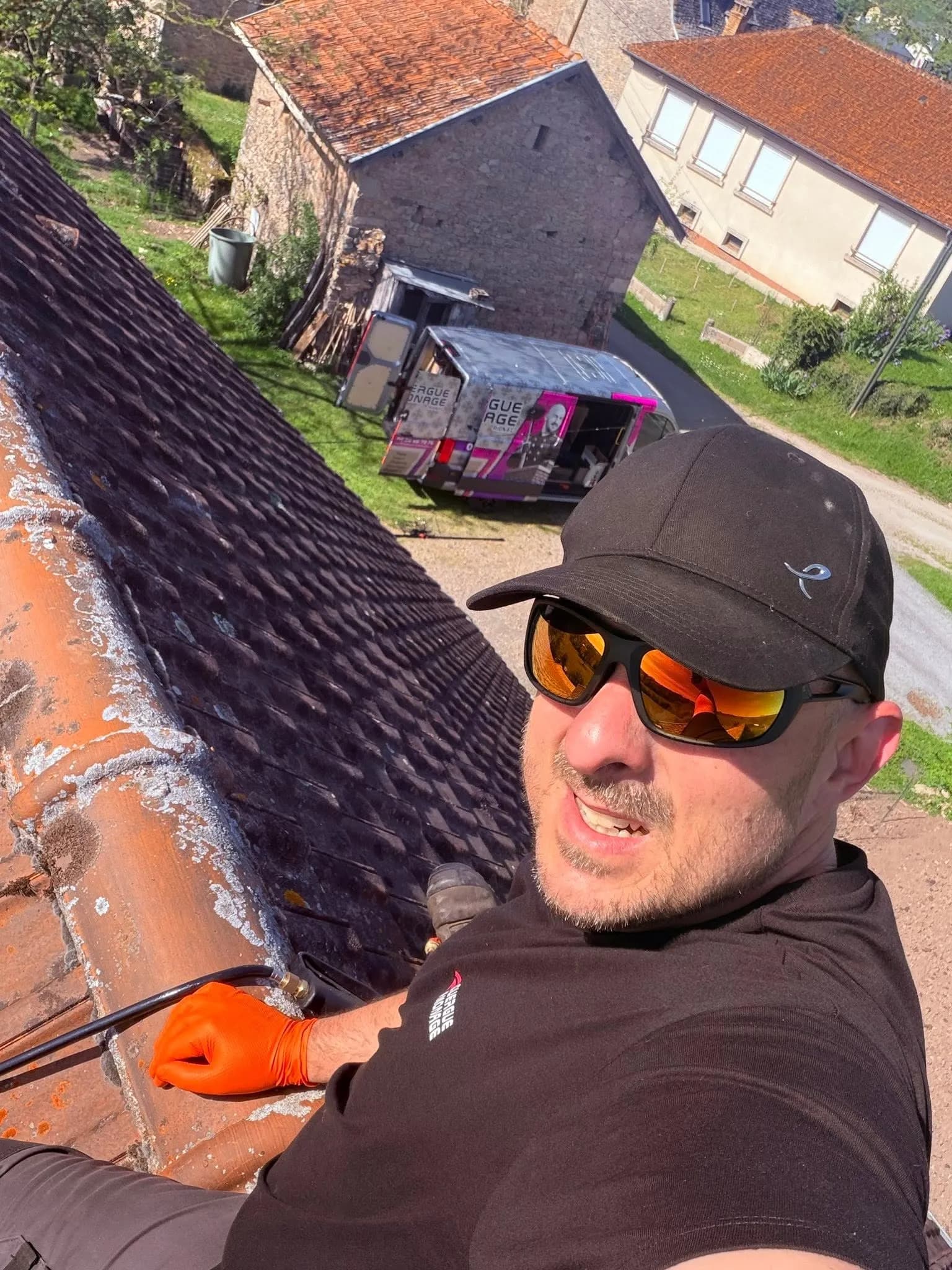 Bartlomiej Lisiewicz en intervention sur un toit, gant de protection orange, camion Rouergue Ramonage visible en contrebas